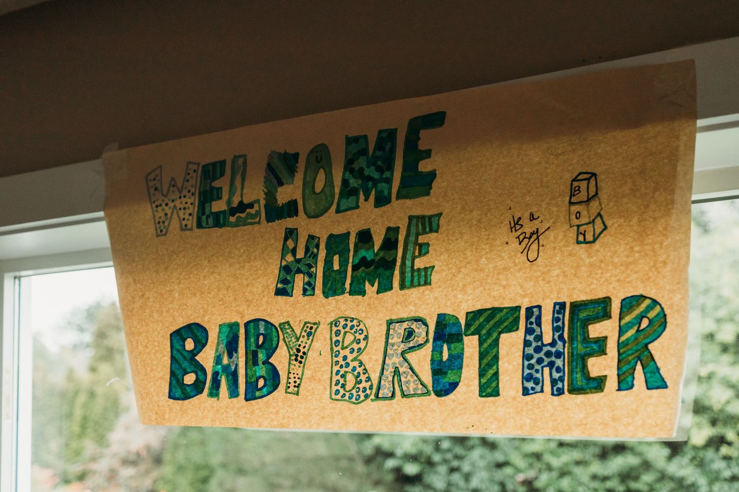 Handmade paper banners with green text spelling out 'In case baby brother' hang across a sunny window.