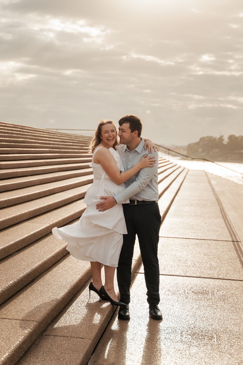 A couple in formal attire hugs romantically on concrete steps at sunrise at the sydney opeara house during their engagement photoshoot