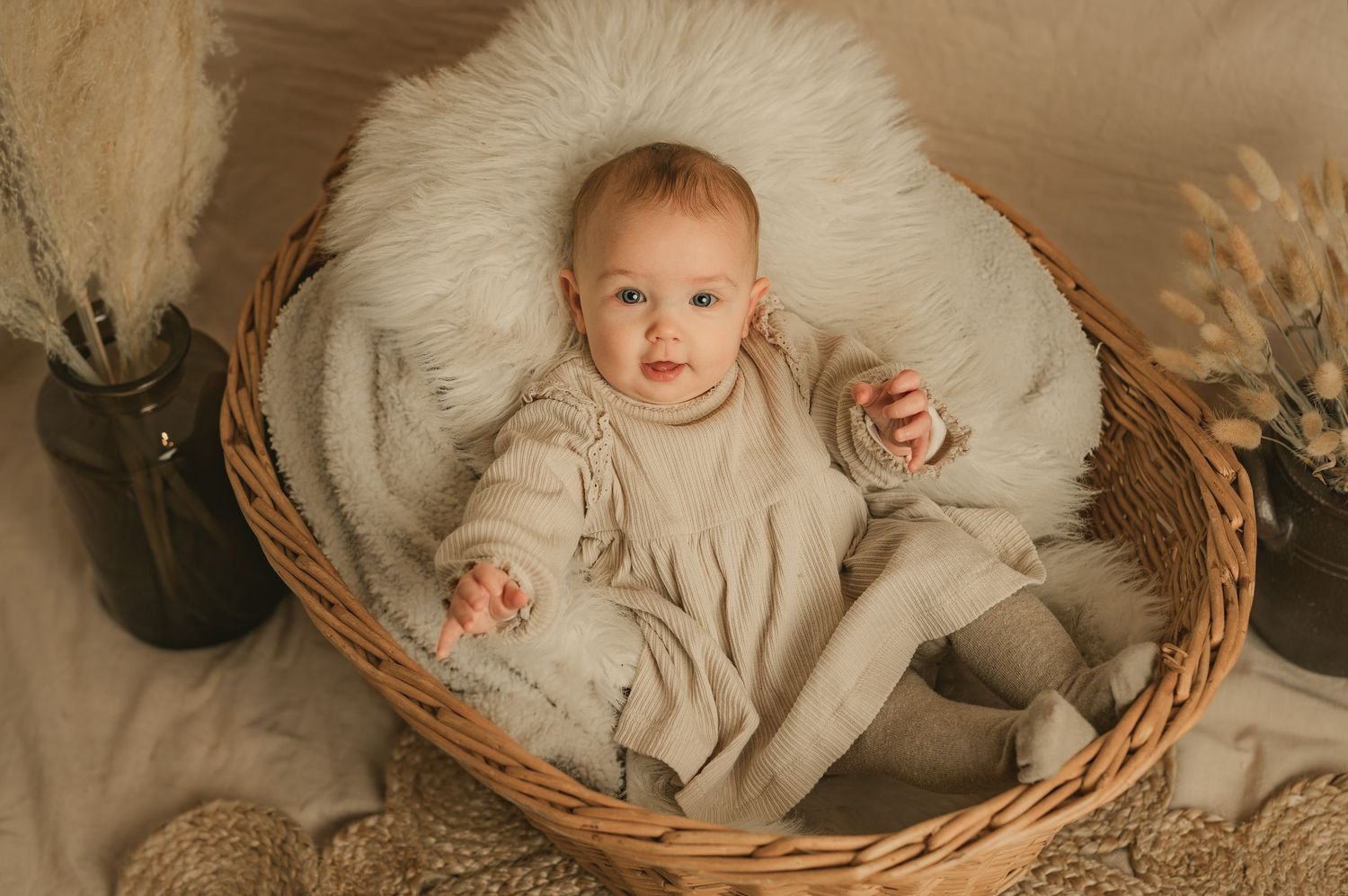 A baby wearing a beige outfit sits in a woven basket with white fur and decorative pampas grass nearby.