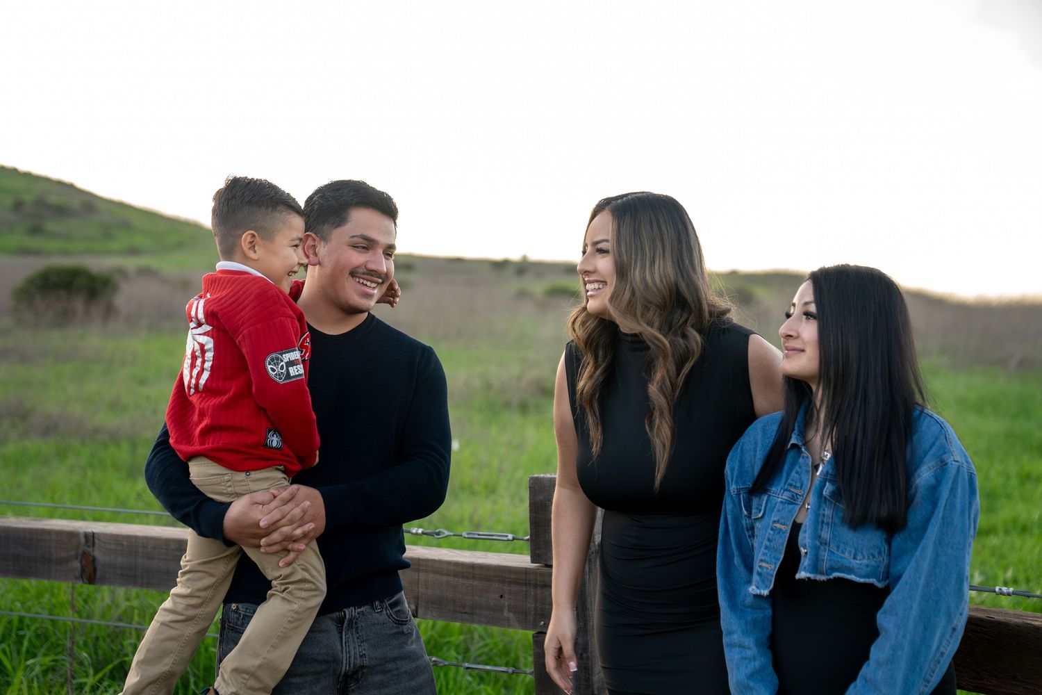 A family enjoys a playful moment together while sitting on a wooden fence in a grassy field.