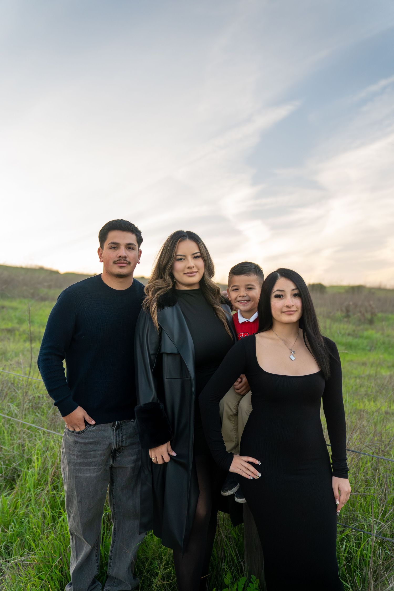 A family of four poses together in a grassy field at sunset with cloudy skies in the background.