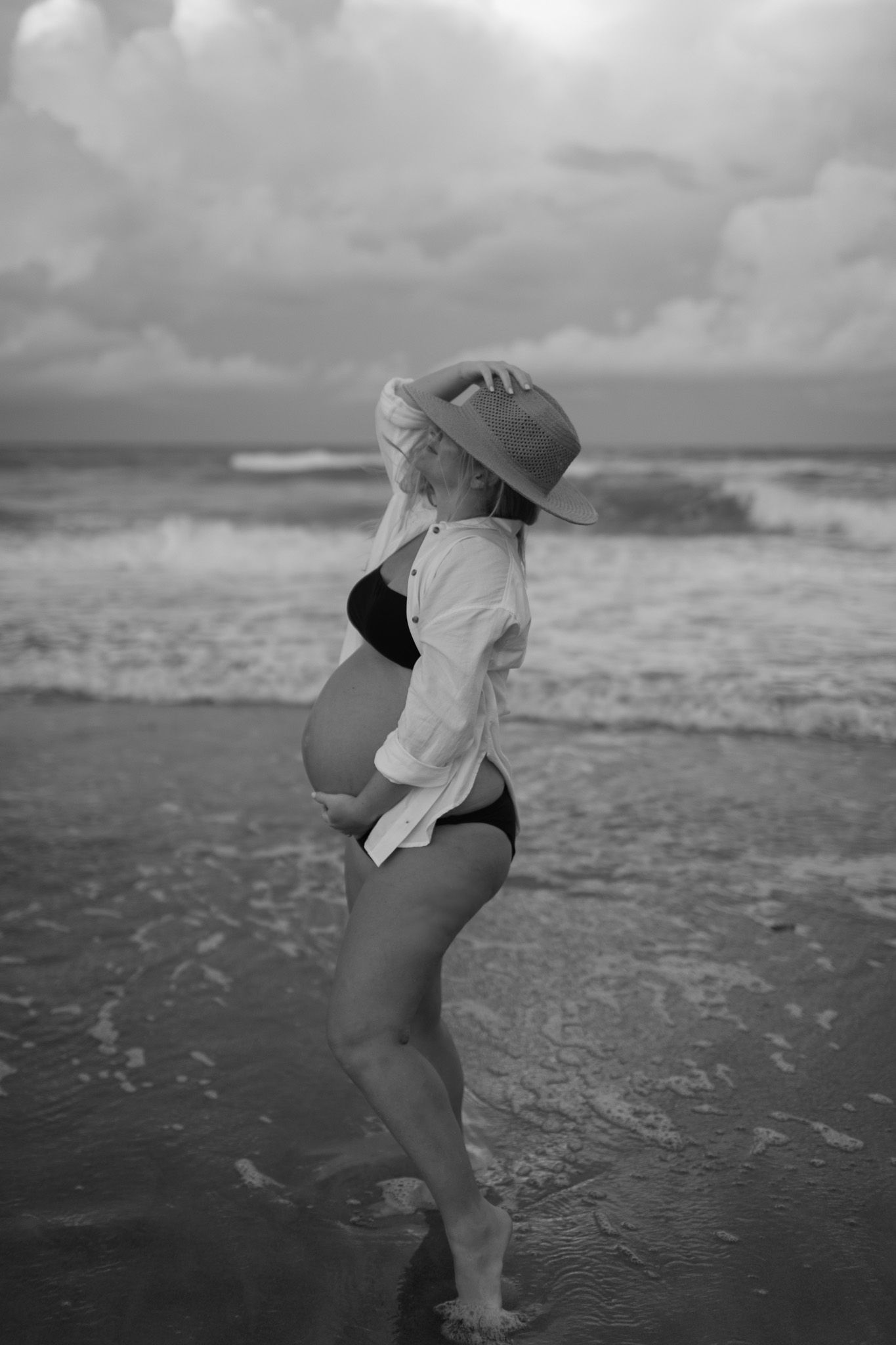 A pregnant woman in a white shirt and straw hat poses outdoors on a moody, overcast sunset on hutchinson island, florida beach