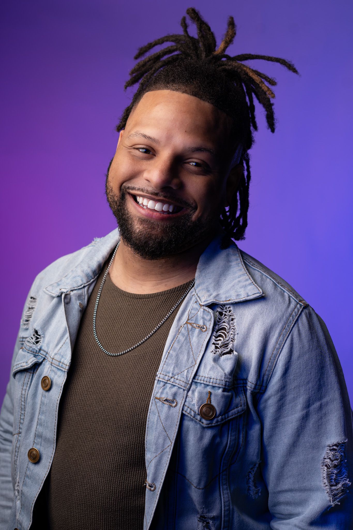 Smiling man with dreadlocks wearing a denim jacket poses against a purple studio background.