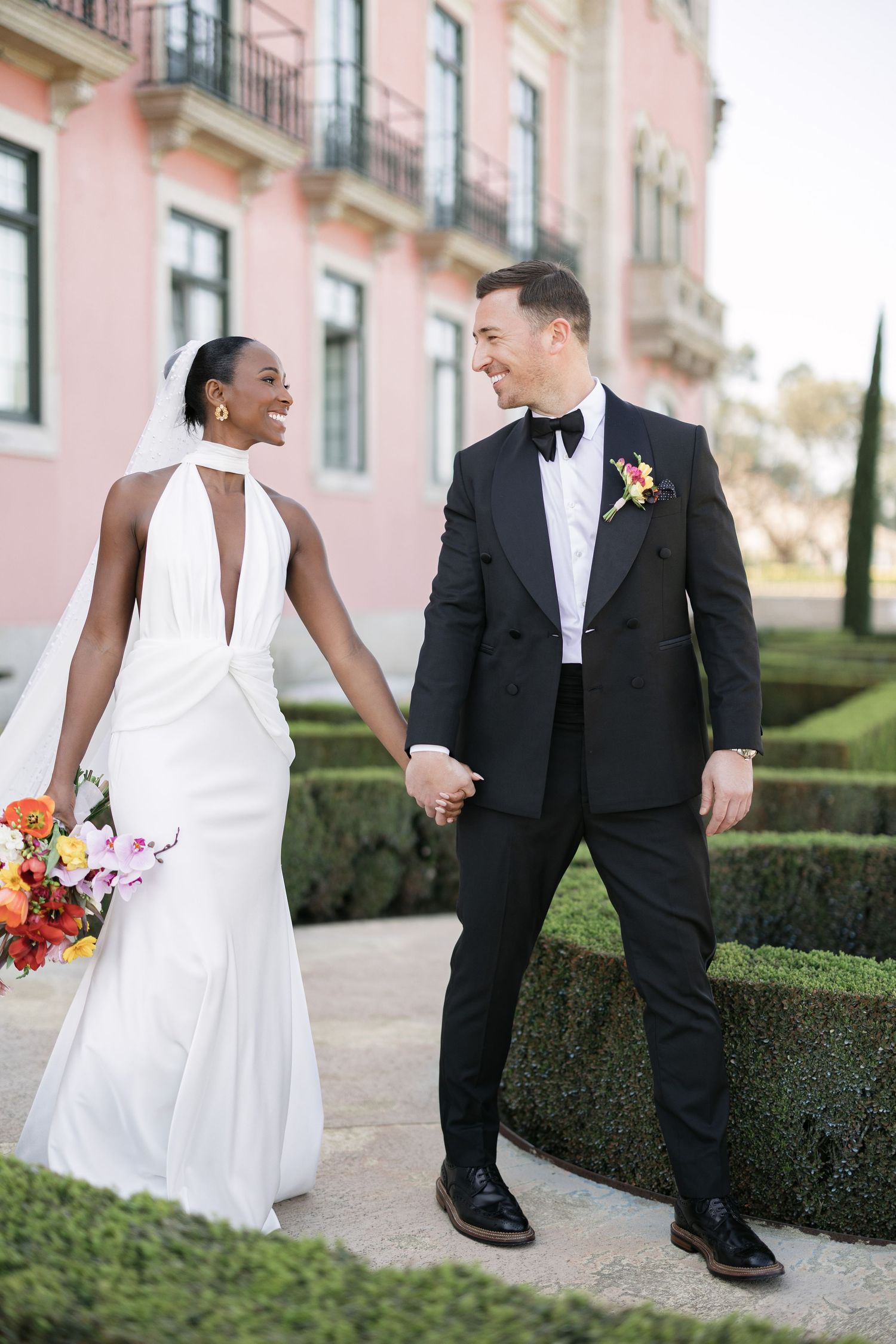 A joyful bride in lace gown embraces groom in black tuxedo with white boutonniere at elegant outdoor wedding.