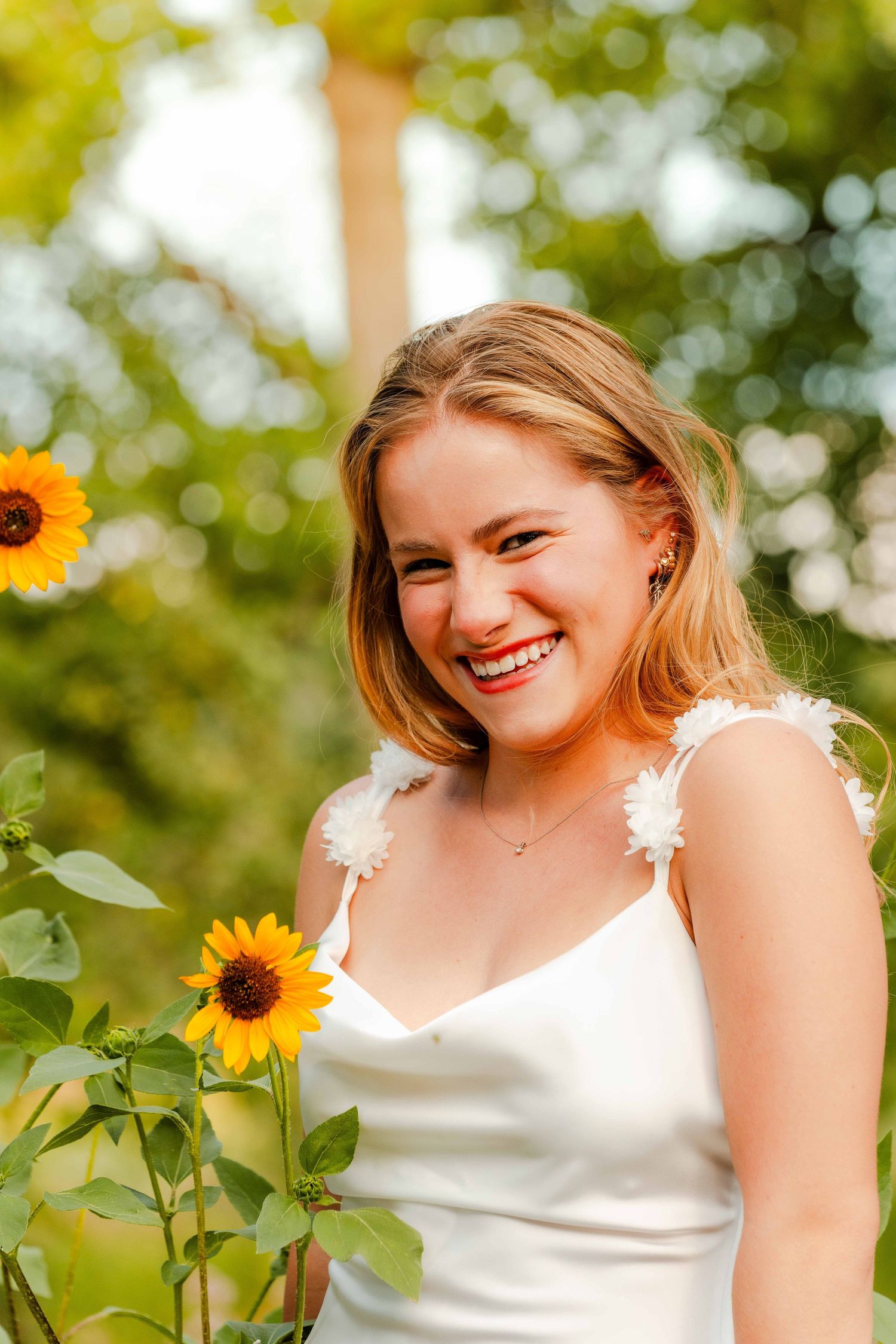 Joyful young woman in a white dress smiles brightly beside vibrant sunflowers in a garden.