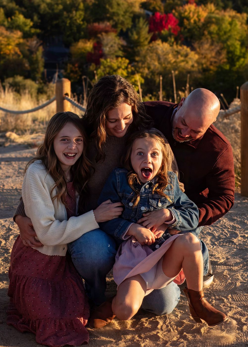 A family shares a joyful moment together while sitting on sandy ground during golden hour with autumn foliage in background.