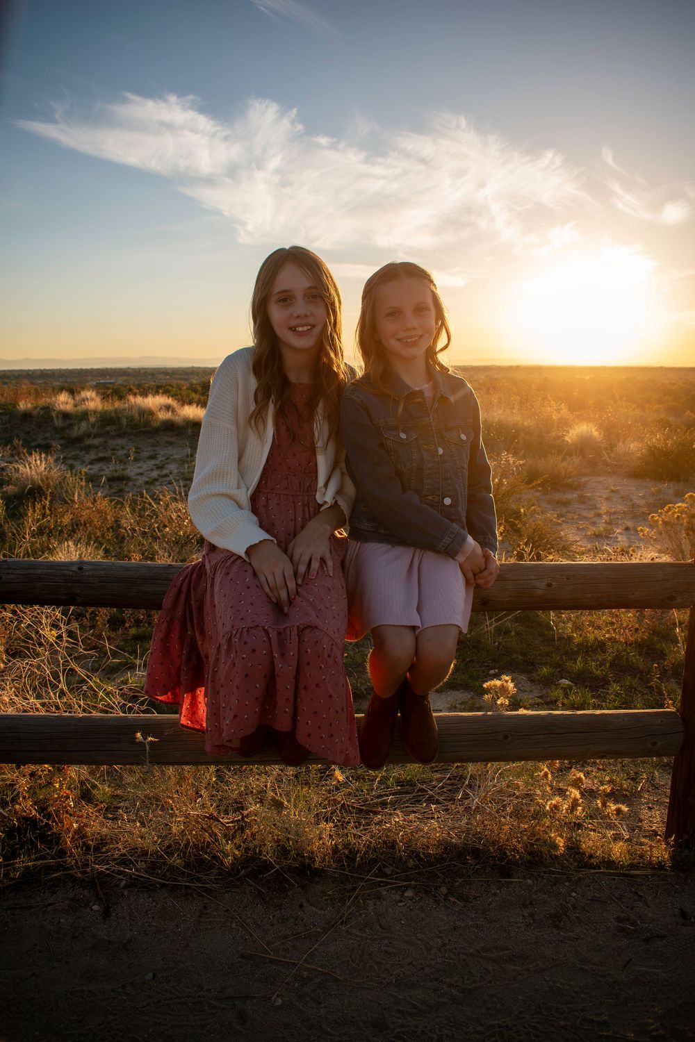 Two sisters sit together on a wooden fence at sunset enjoying the golden hour view over a scenic landscape.