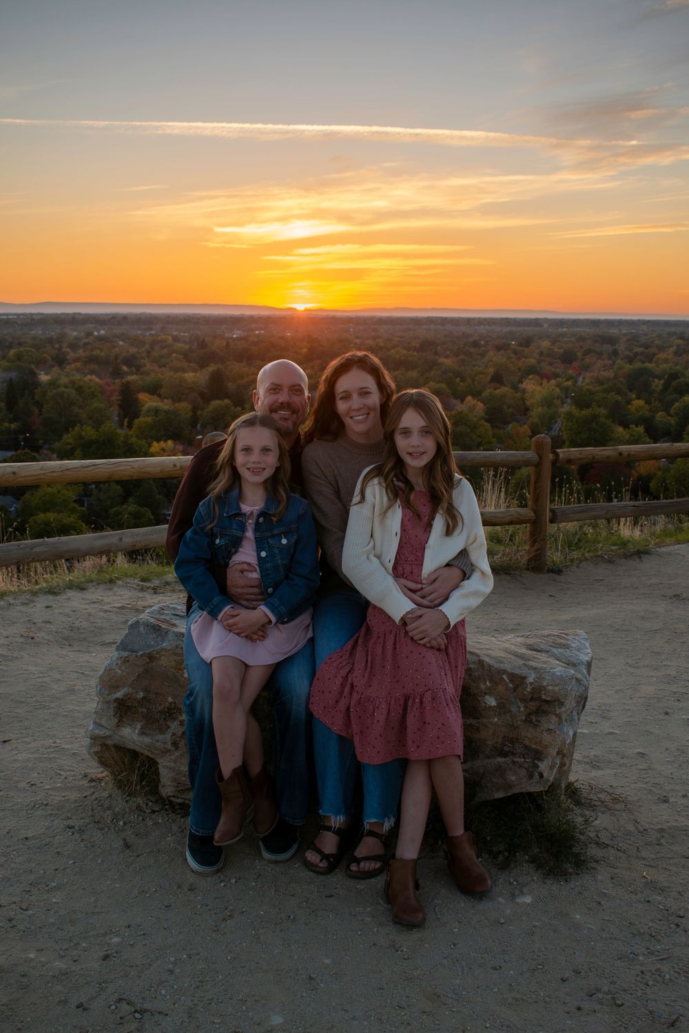 Family sits together on stone wall overlooking scenic autumn landscape at sunset.