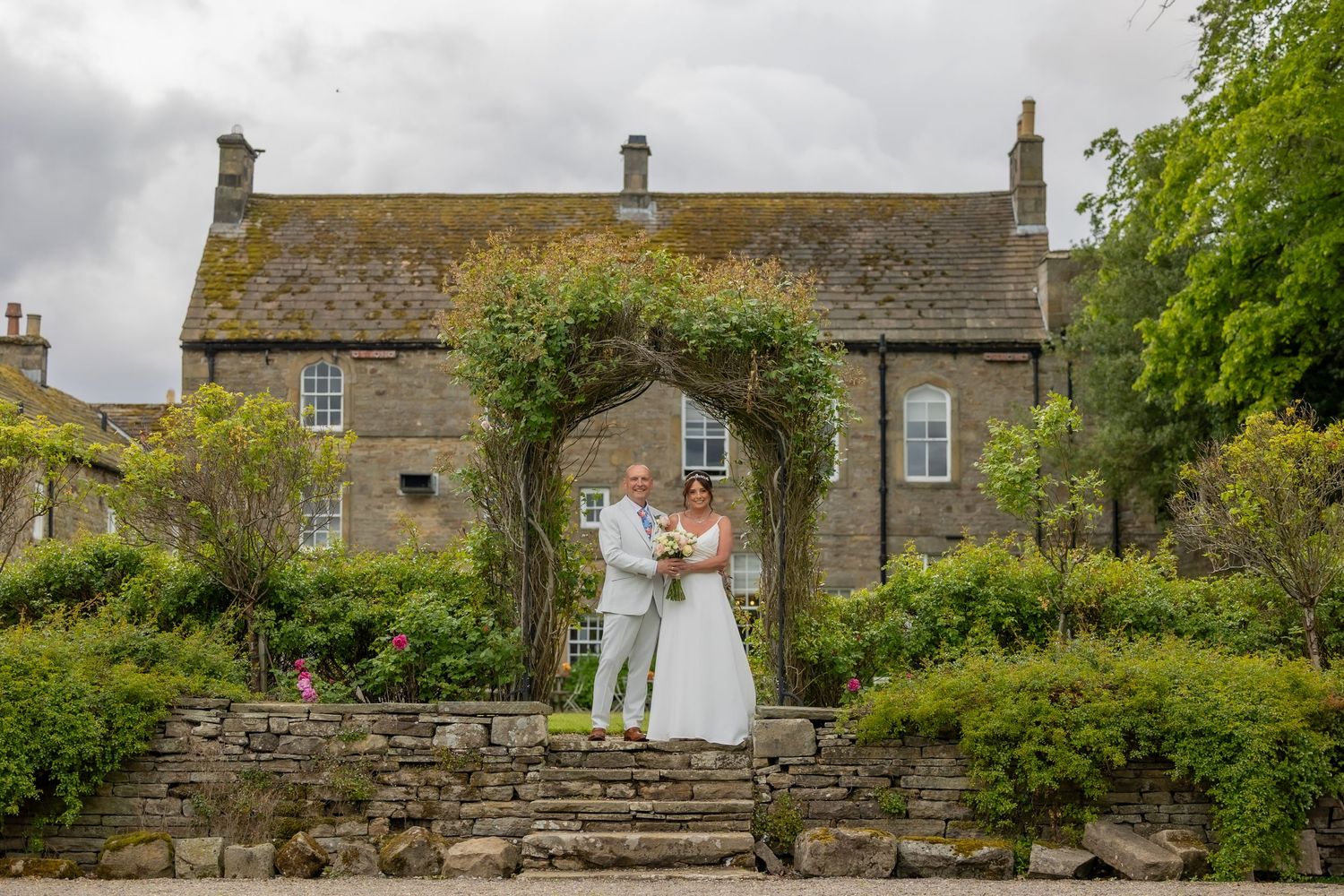 Stone buildings and walls line a path through a traditional Northumberland village with lush green foliage. Lord Crewe Arms Wedding