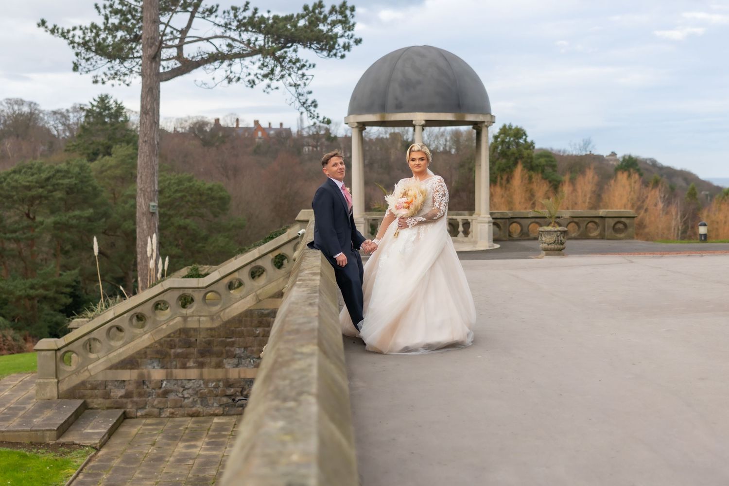 A couple in wedding attire stands together on a stone bridge surrounded by autumn foliage in a scenic outdoor setting. Rushpool Hall Wedding