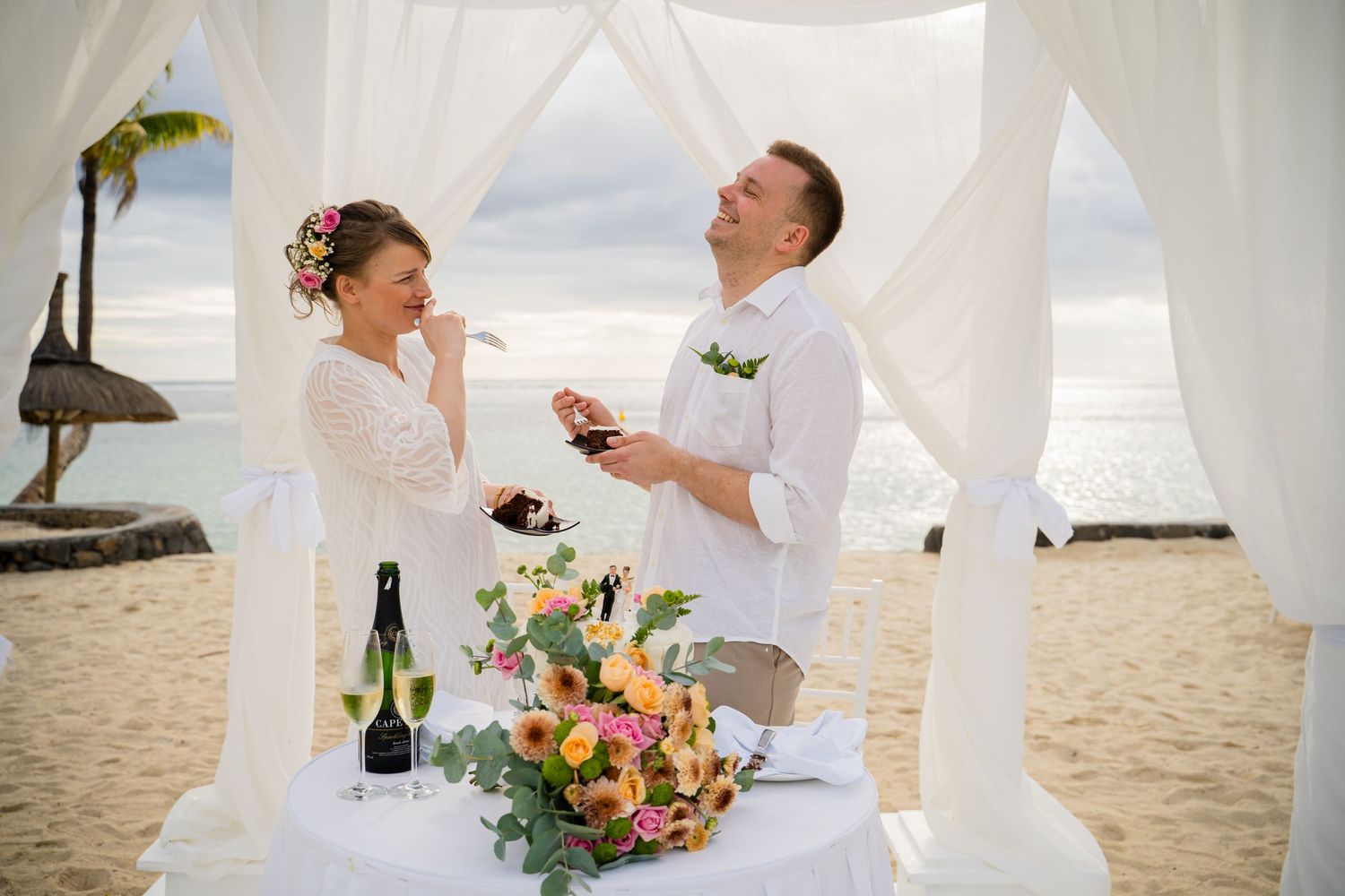 Romantic beach wedding ceremony with white fabric draping and tropical floral arrangements on a sandy shore. Shot by Luminair Studio