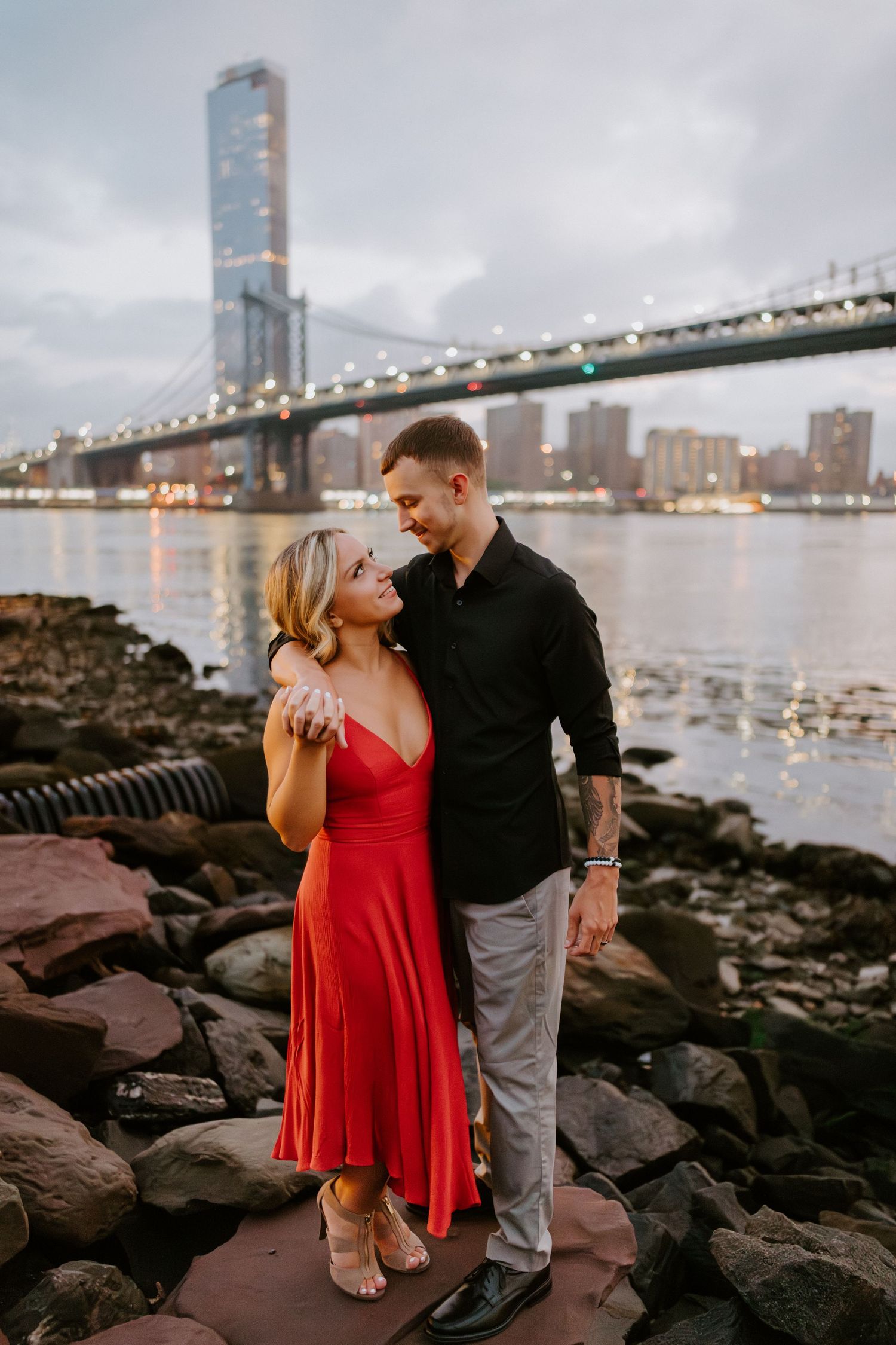 Couple embraces near rocky shoreline with Manhattan Bridge and NYC skyline at dusk.