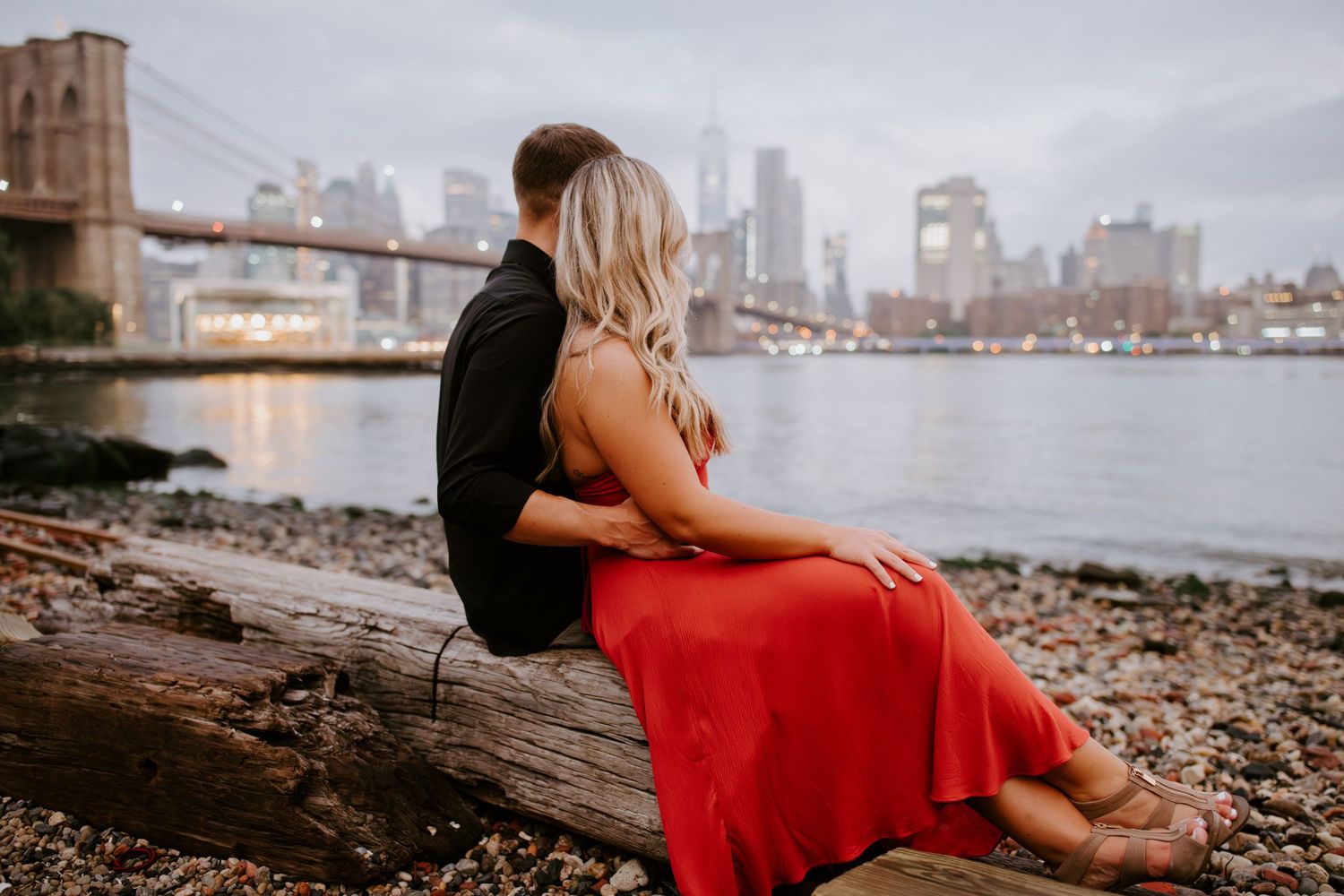 Couple sits on driftwood log with Brooklyn Bridge and New York City skyline behind them.