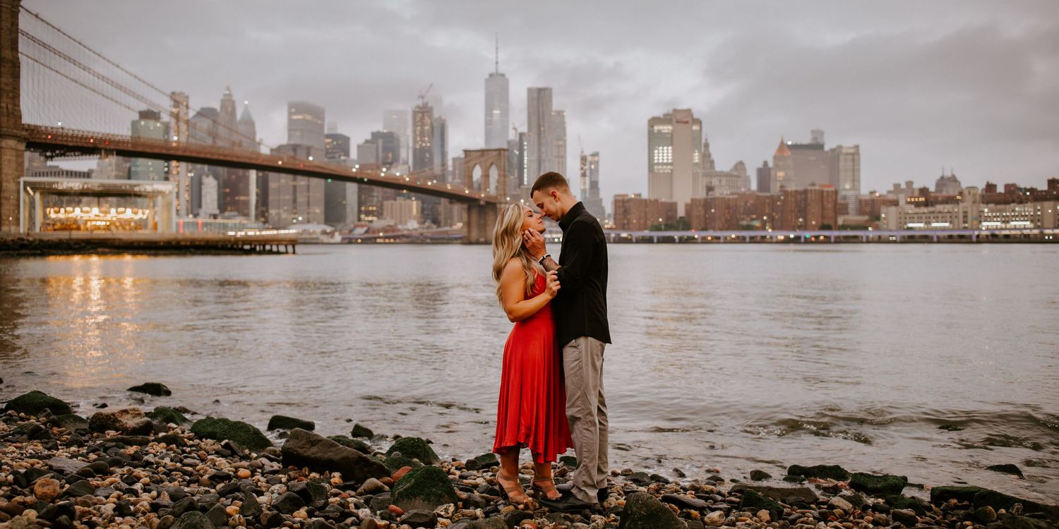 Couple shares a tender kiss by the water with Brooklyn Bridge and NYC skyline backdrop.