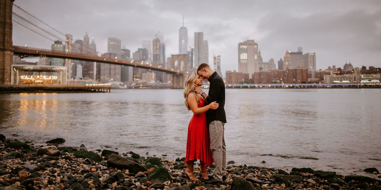Couple embraces romantically on rocky shore with Brooklyn Bridge and Manhattan skyline at dusk.