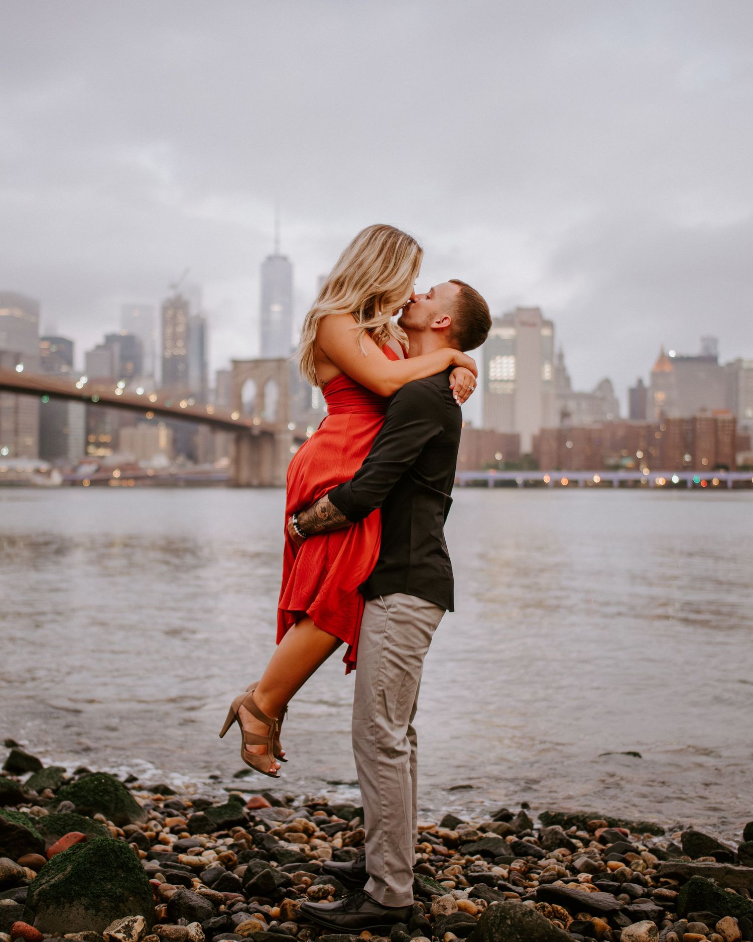 Man lifts woman in red dress for a kiss with Brooklyn Bridge and NYC skyline behind them.