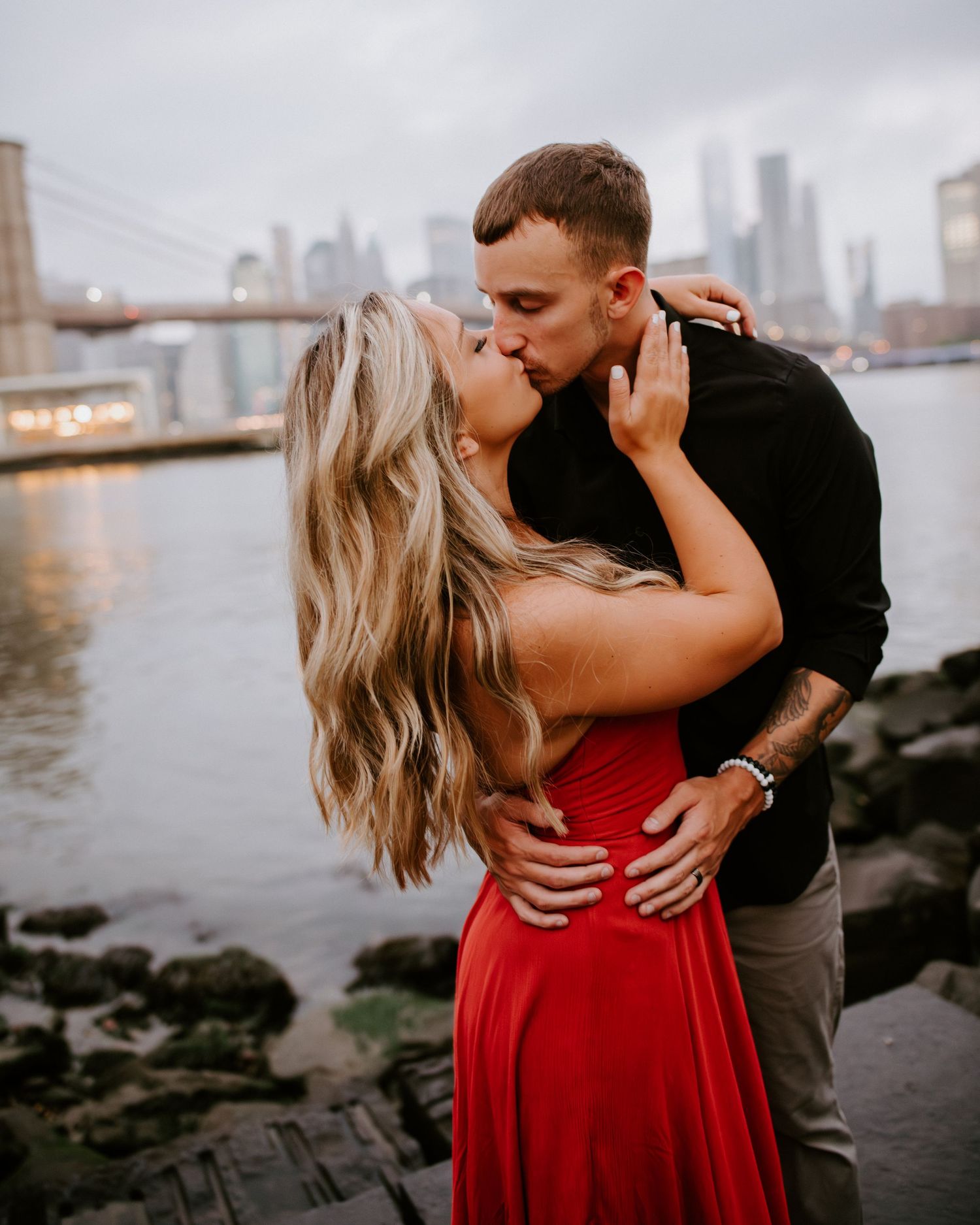 Couple sharing a romantic kiss near the Brooklyn Bridge with NYC skyline in the background.