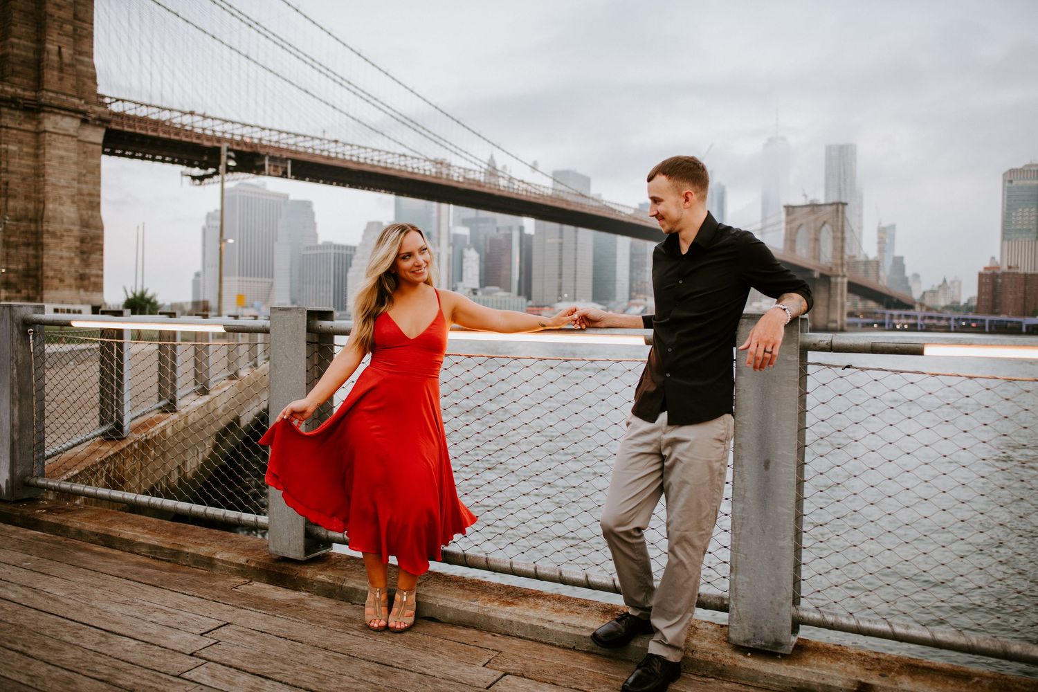 Woman in red dress dancing with man on Brooklyn Bridge waterfront promenade.