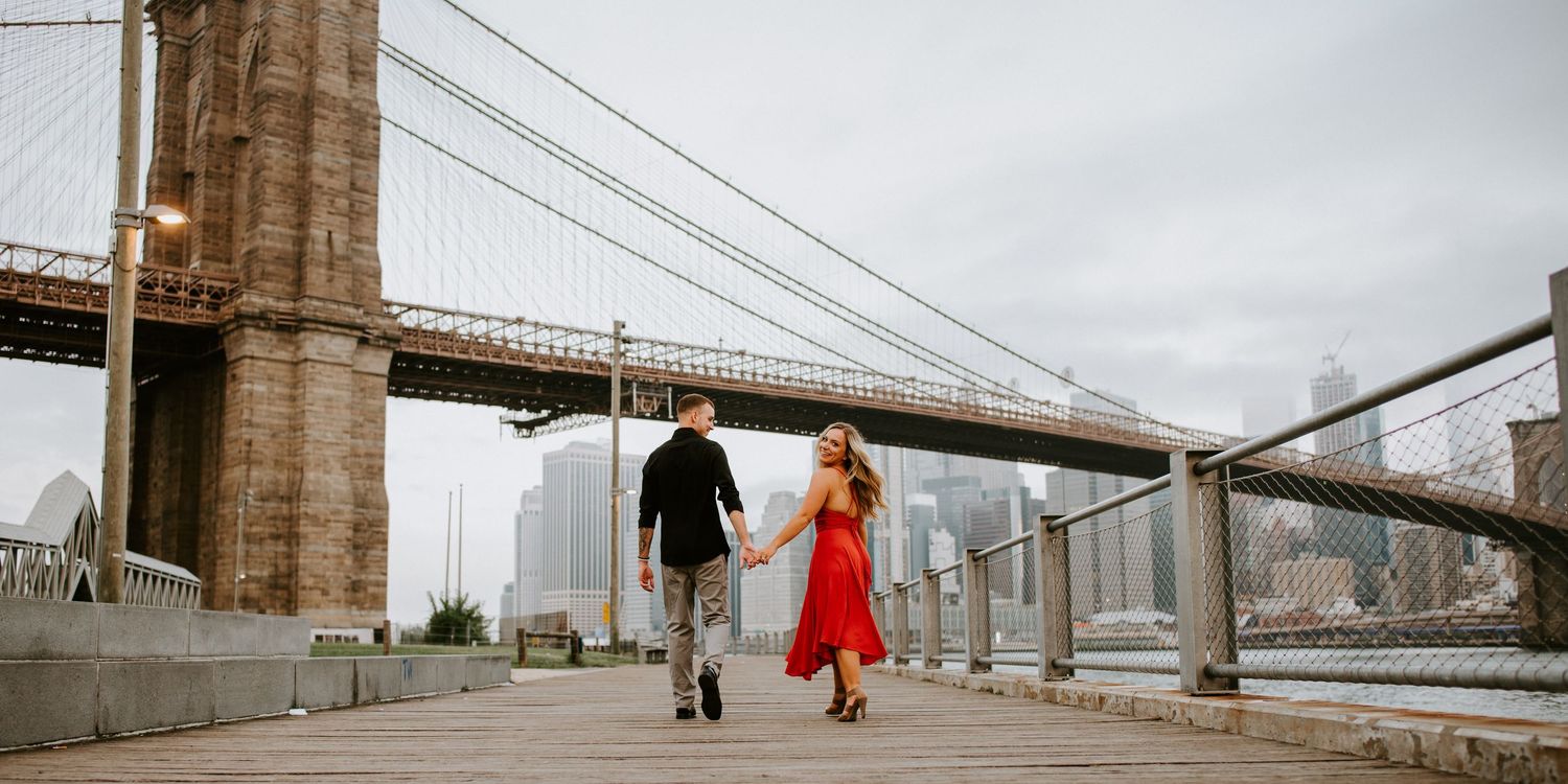 Couple holding hands walking along Brooklyn Bridge park pathway with city skyline.
