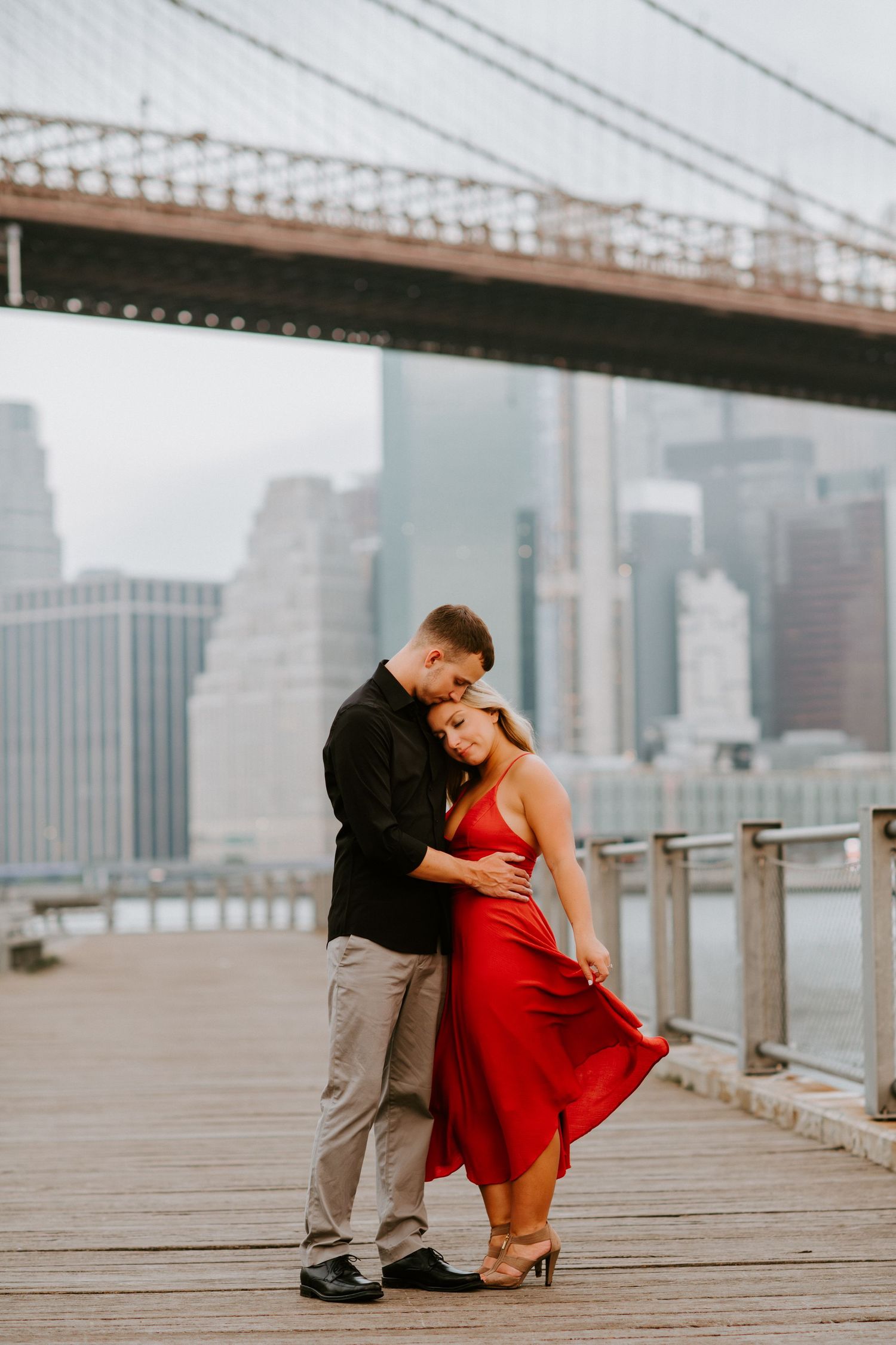Couple embracing on wooden boardwalk with Brooklyn Bridge and Manhattan skyline behind them.