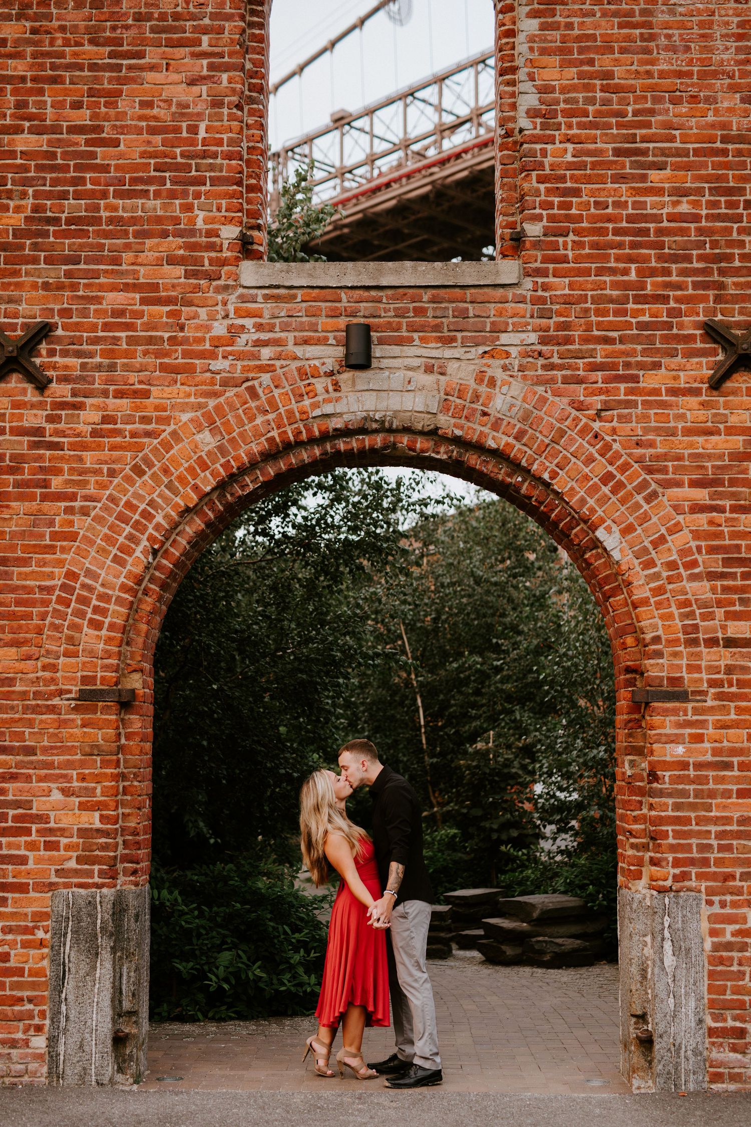 Couple holding hands and nearly kissing under a rustic red brick archway near Brooklyn Bridge.
