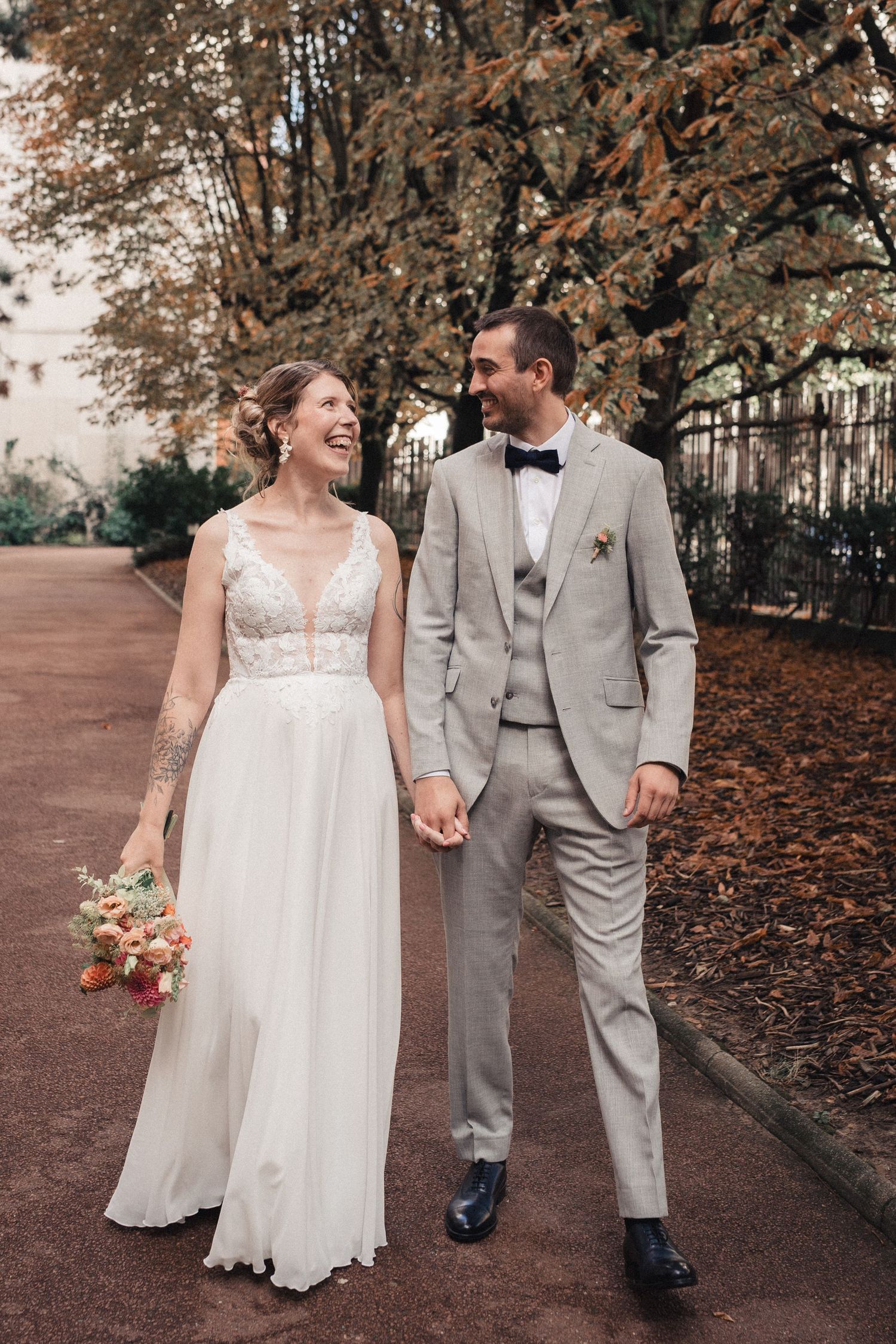 A couple in wedding attire shares a joyful moment while walking along a path covered in autumn leaves.