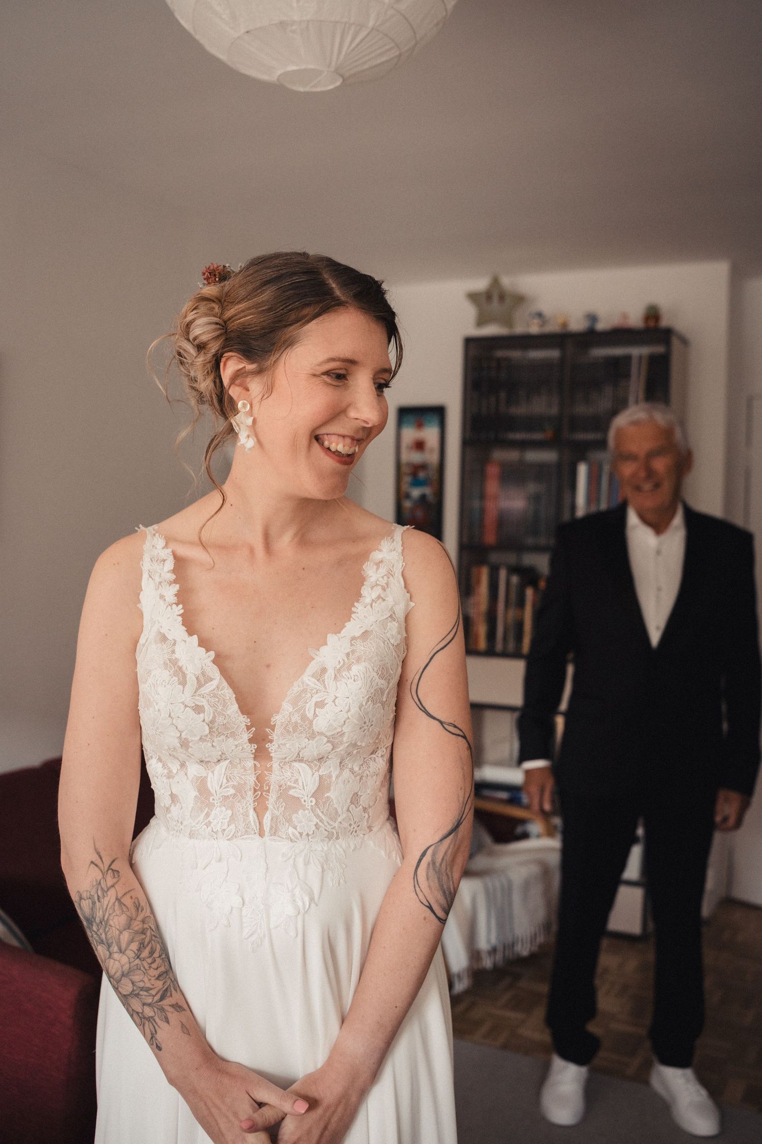 A bride in a white lace wedding dress with tattoos smiles joyfully while getting ready with someone in formal attire behind her.