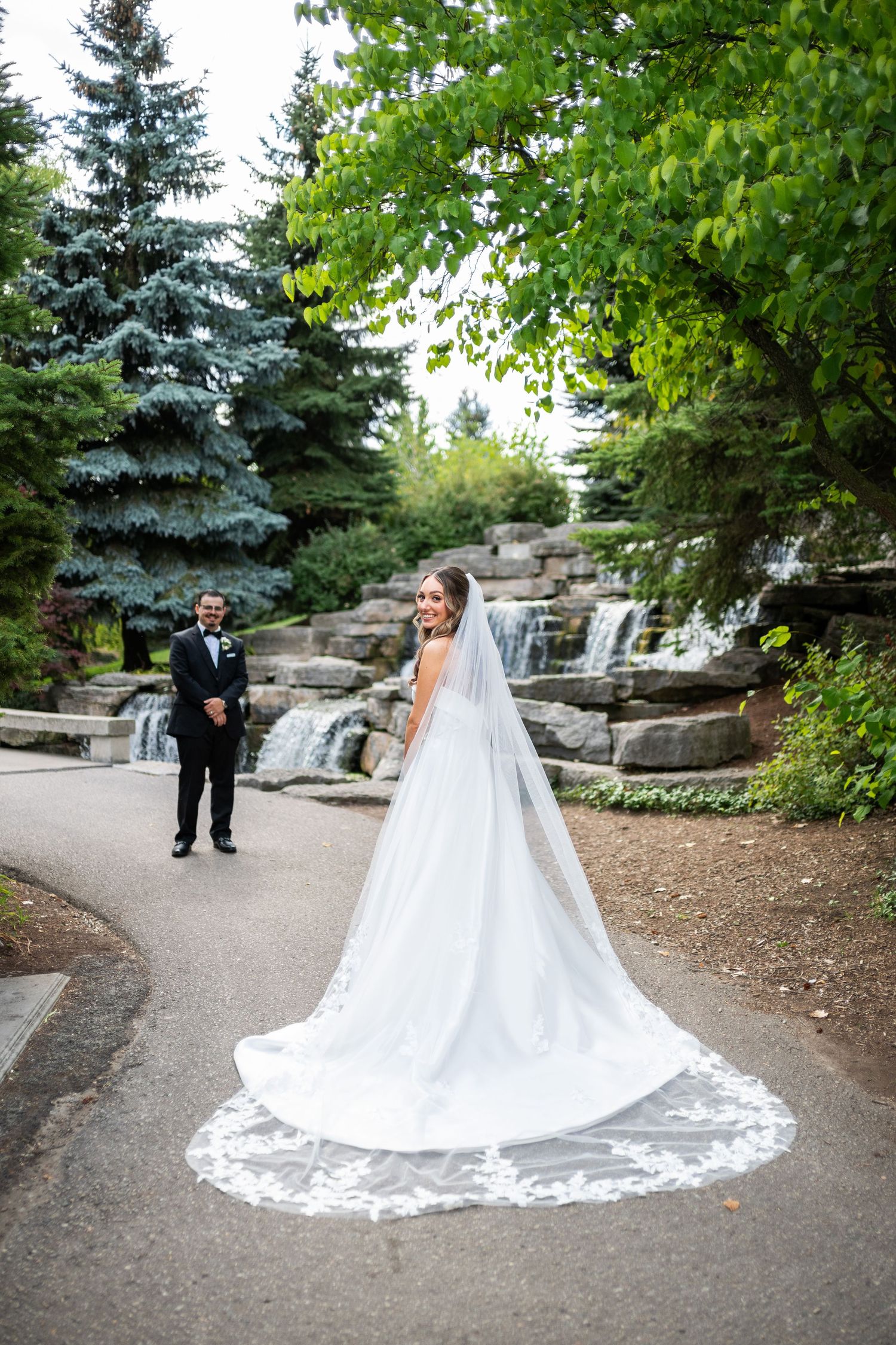 A long flowing white wedding dress with cathedral train displayed on a garden path surrounded by evergreen trees.