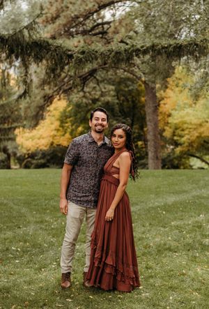 A couple poses together in a park during autumn, wearing casual attire against a backdrop of trees and grass.