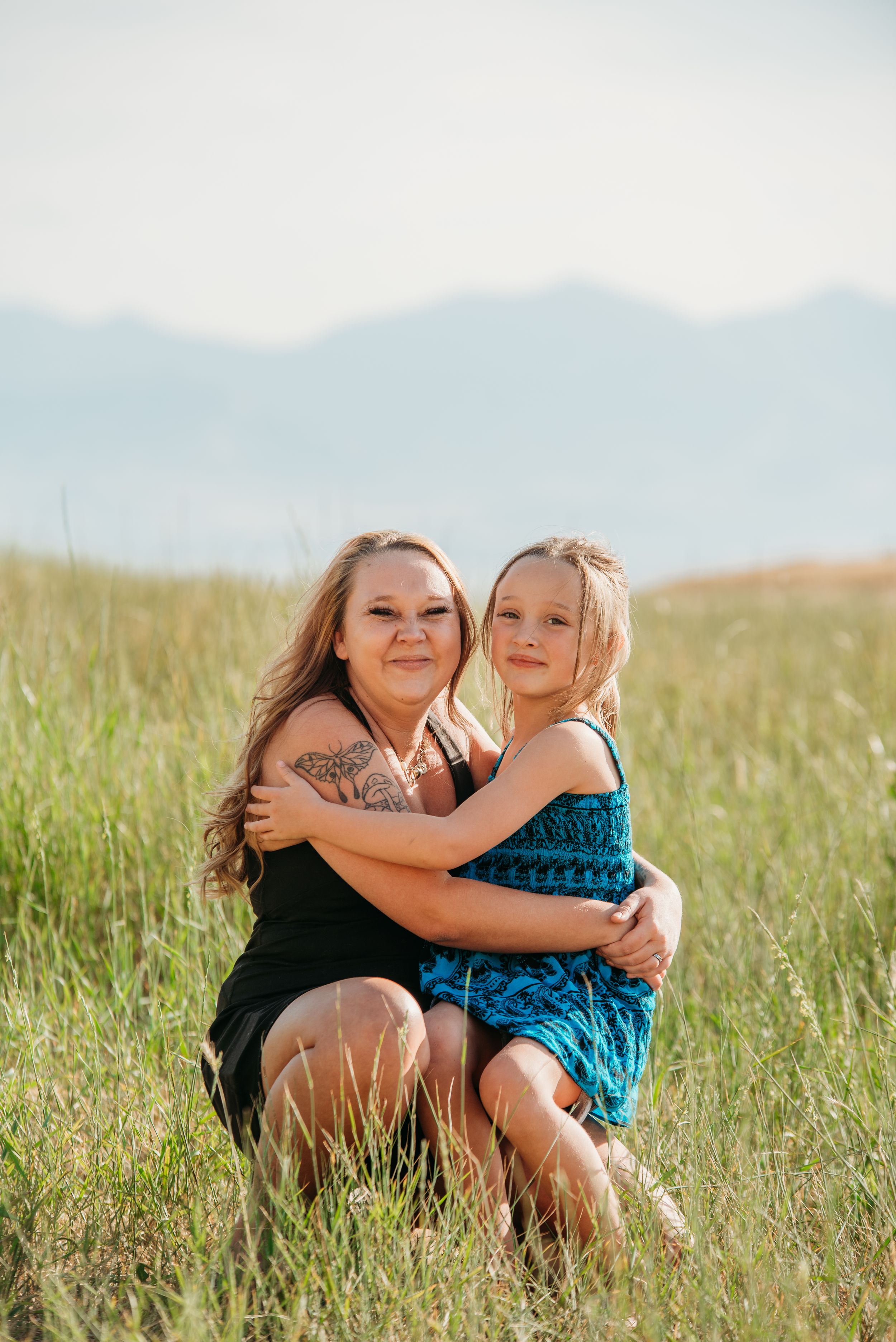 Two people embrace while sitting in a grassy meadow with mountains visible in the misty background.