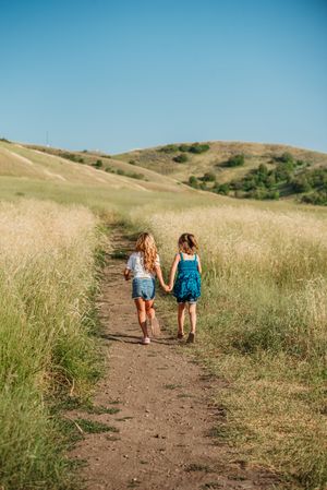 Three people walk along a dirt trail through golden grass on rolling hills under bright blue sky.