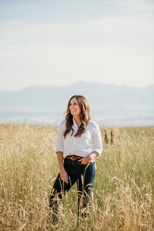 Person in a white button-down shirt and jeans stands in a golden wheat field with mountains in the background.
