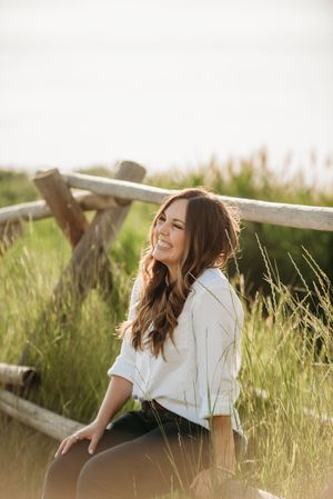 Person in white shirt sits by wooden fence in tall grass at sunset, smiling and laughing with carefree expression.