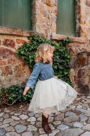 A child in a denim top and tulle skirt twirls on a cobblestone path against a stone wall with climbing ivy.