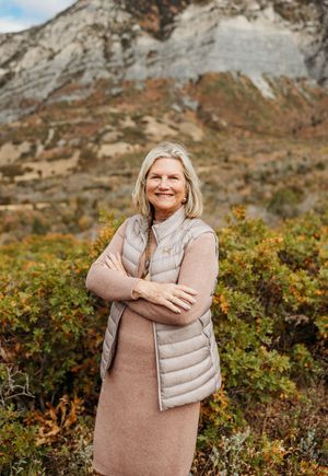 A person wearing a beige sweater and vest poses outdoors against a scenic mountain backdrop with autumn foliage.