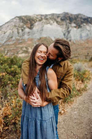 A couple shares a tender moment along a mountain trail, with rugged peaks and autumn foliage in the background.