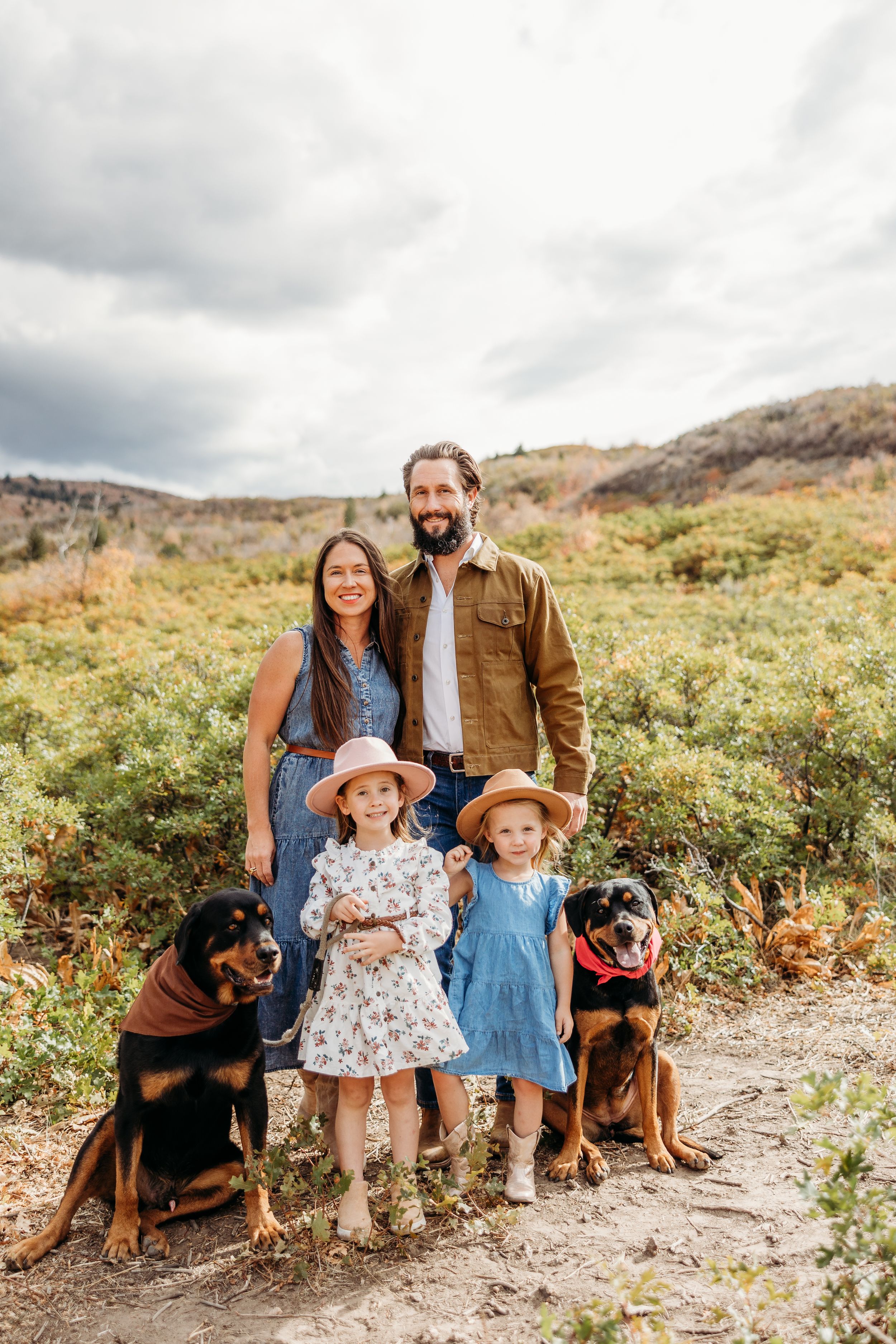 Family with two children and two dogs posing together on a scenic mountain trail on a cloudy day.