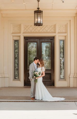 A romantic wedding couple embraces under an elegant colonnade with ornate architectural details and hanging lanterns.