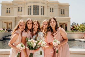 Bridesmaids in coral dresses pose together with bouquets outside a grand stone building.
