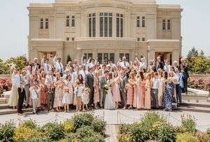 Large wedding party poses together outside a grand stone church building in varying shades of pink and peach attire.