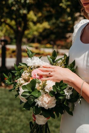 A delicate bridal bouquet with white peonies, pink roses and greenery held by hands wearing an elegant bracelet.