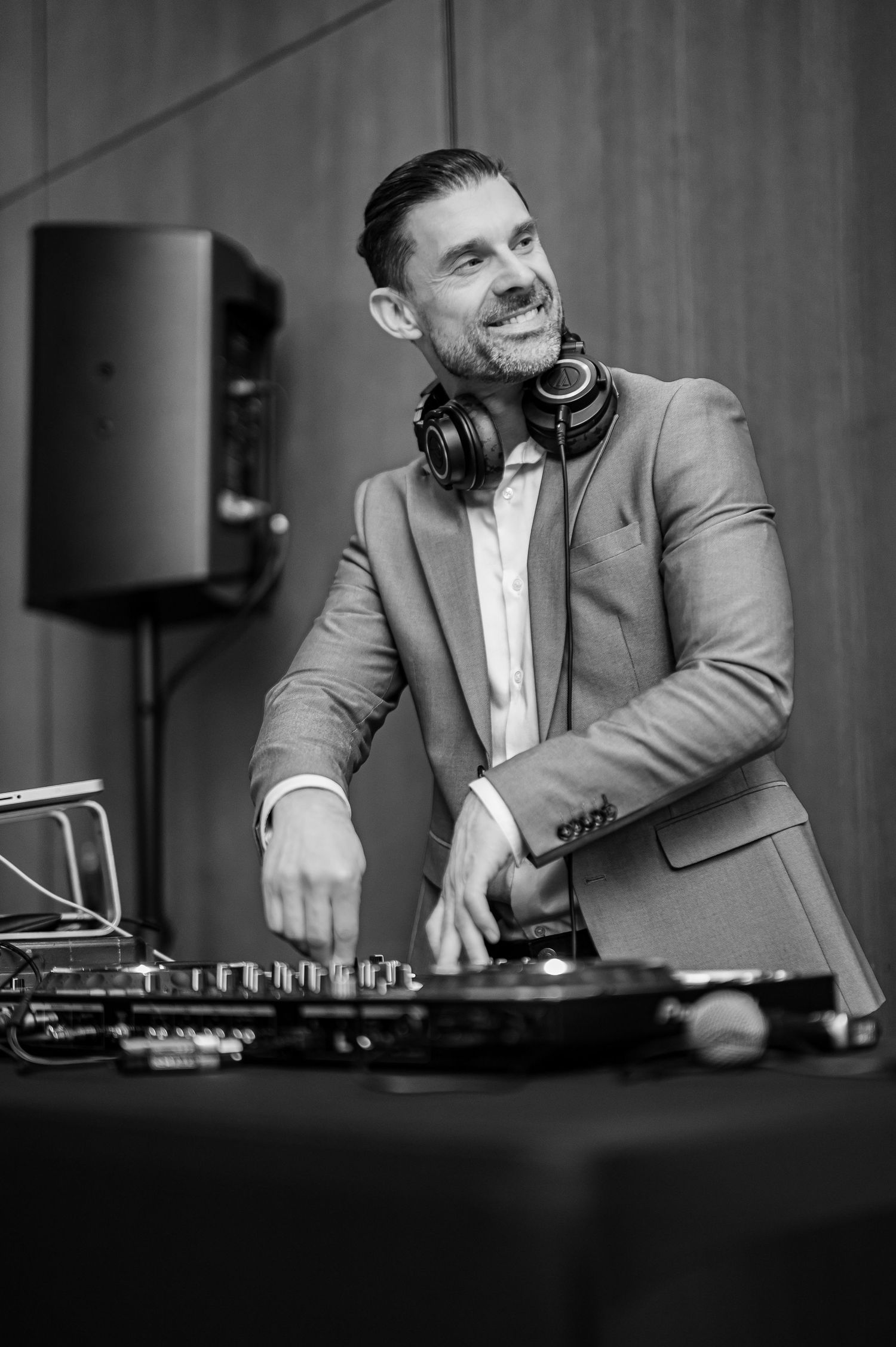 Professional DJ in gray suit jacket and headphones performs at mixing deck in black and white photograph.