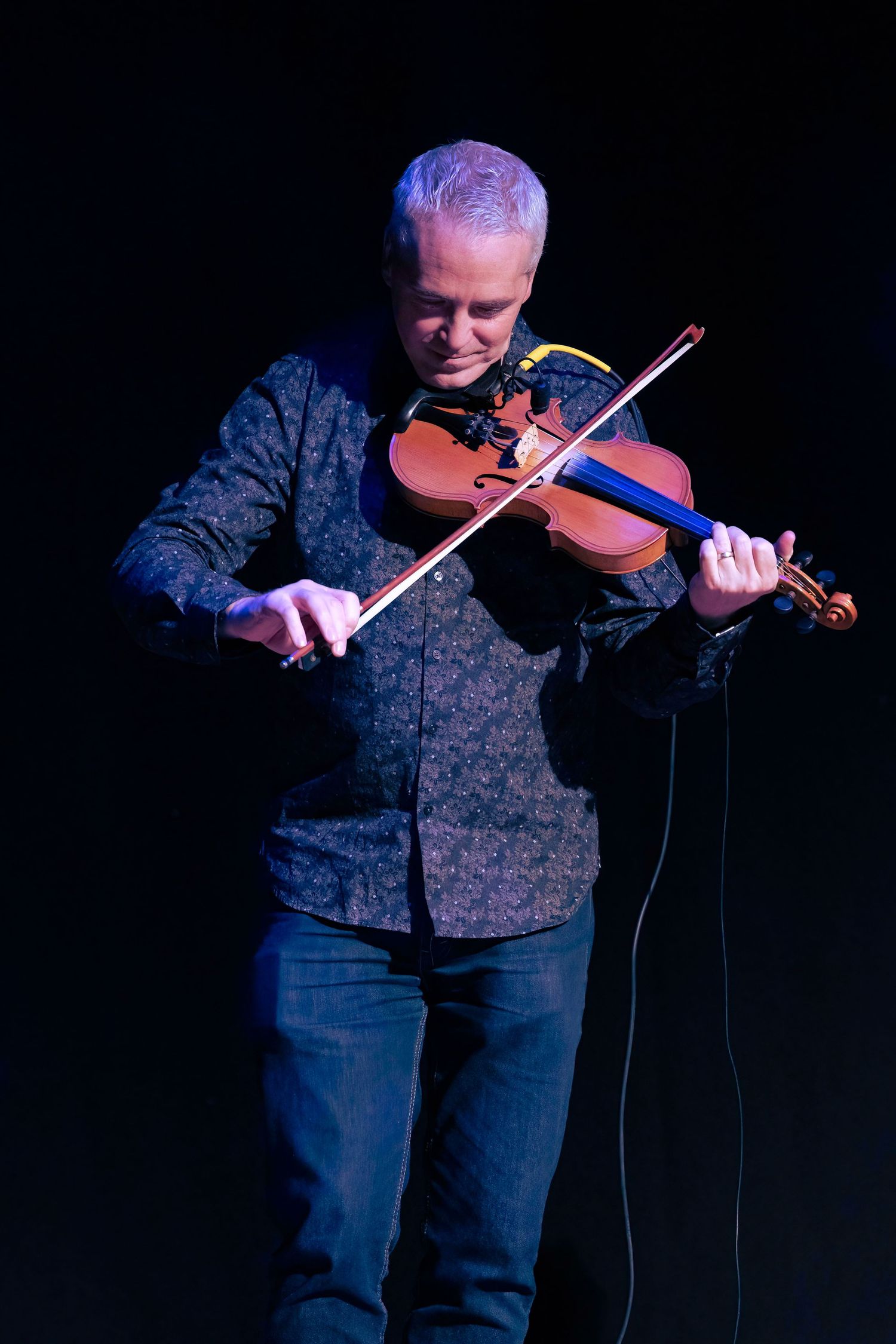 A violinist performs on stage in a dark setting wearing a patterned shirt and jeans.