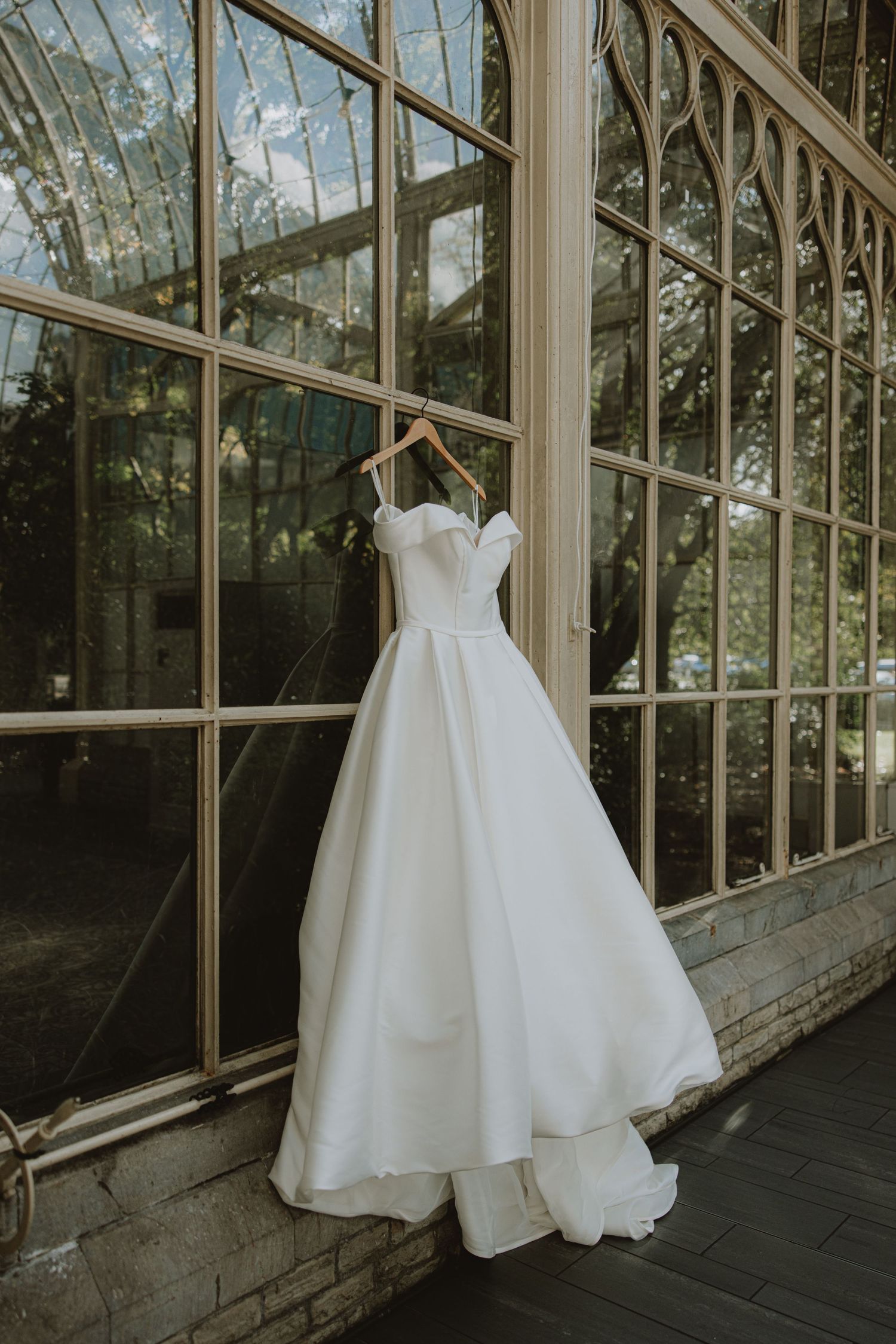 Four elegant white wedding dresses displayed in sequence against glass windows of a vintage greenhouse conservatory.