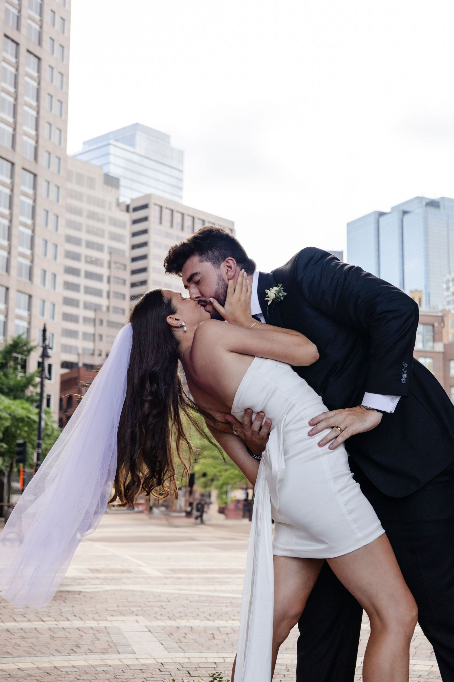 A romantic dip kiss between newlyweds in formal wedding attire on a city plaza with tall buildings in the background.