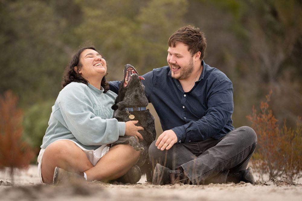 A couple photo with their Cattle Dog in the middle. Sitting on a sandy path and laughing at a dog photoshoot.