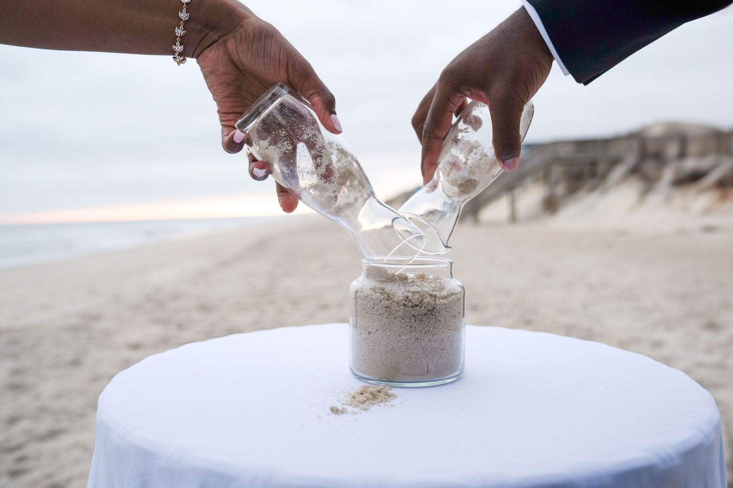 Two hands pouring sand into a glass jar during a beach ceremony ritual.
