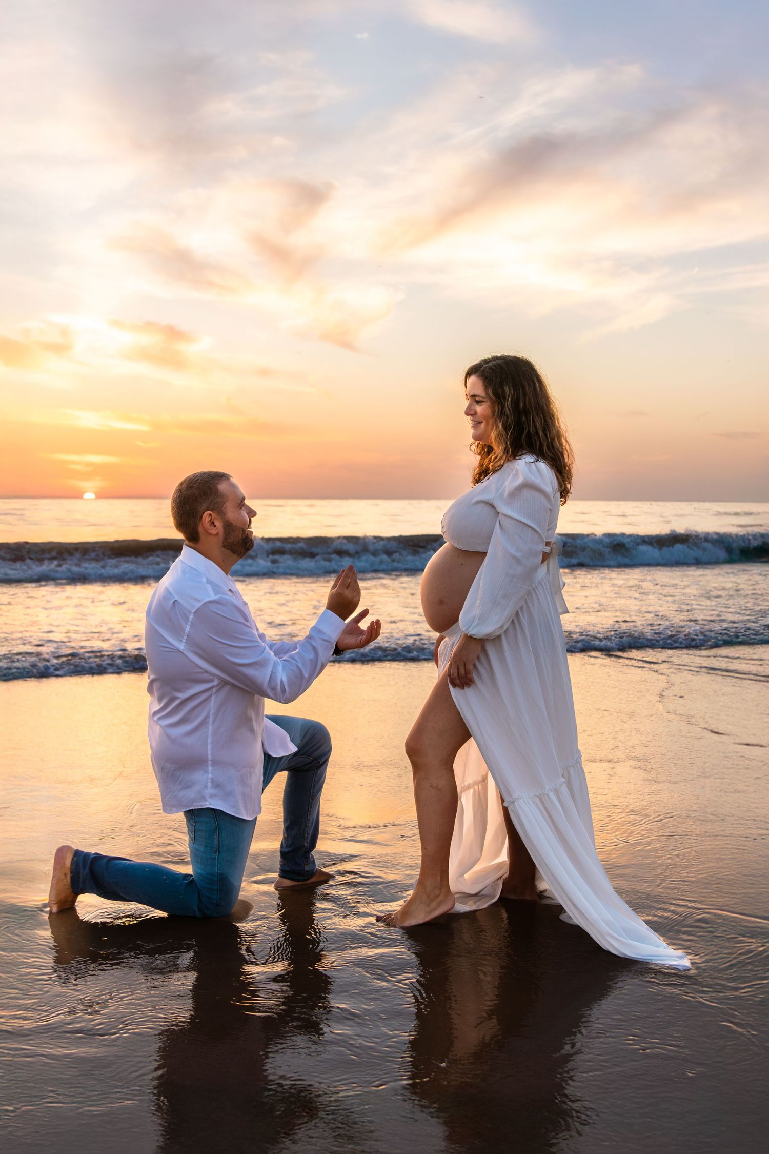 Couple sharing intimate moment on beach at sunset with waves and orange sky in background.