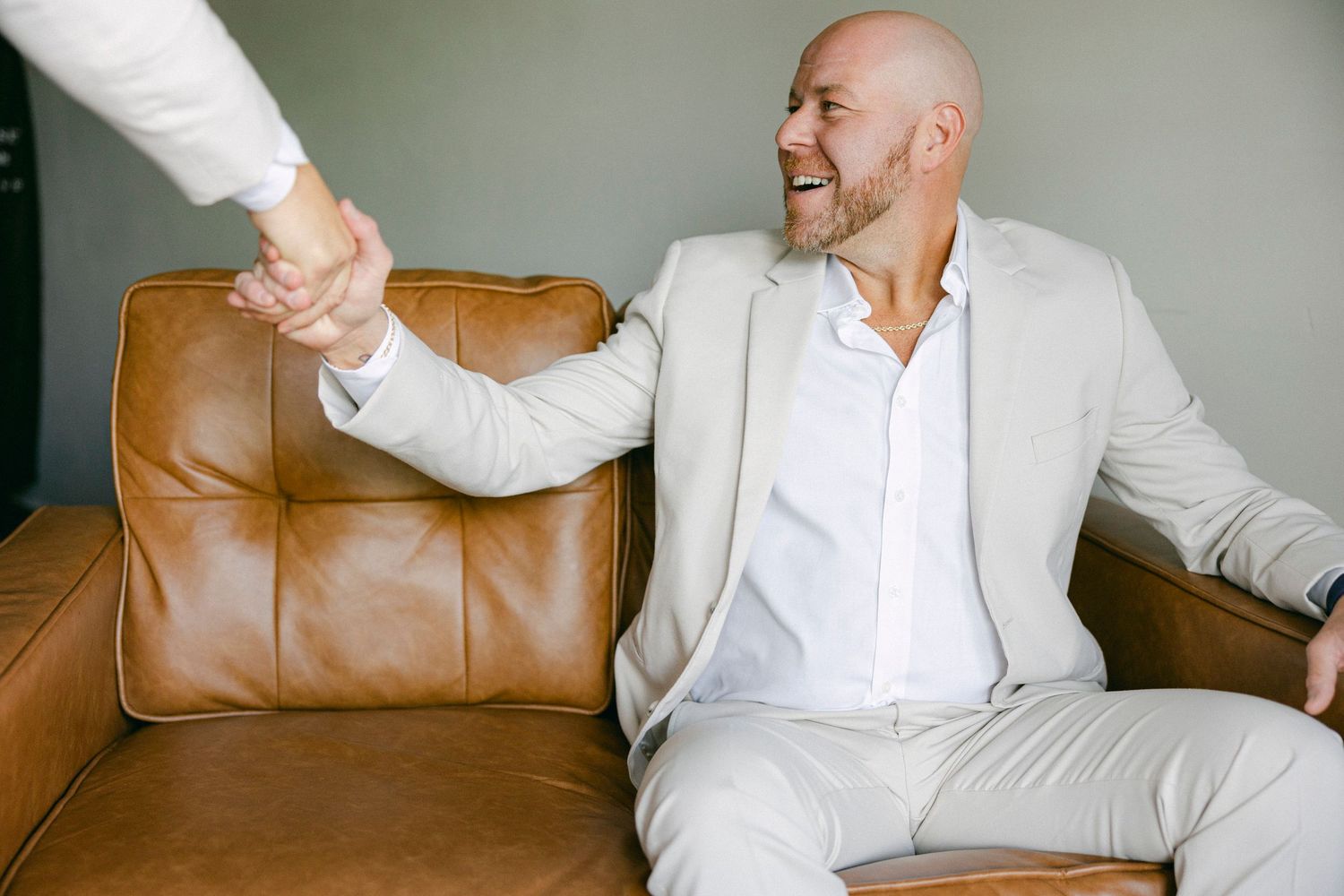 A person in a white shirt sits on a brown leather couch while gesturing and laughing during a conversation.
