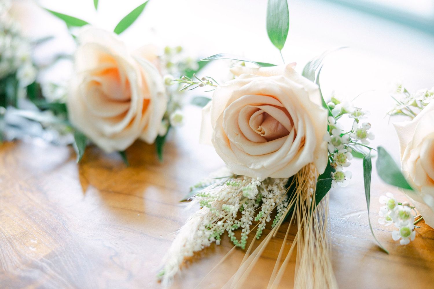 Delicate peach roses with greenery and wheat stalks arranged on wooden surface for wedding decor.